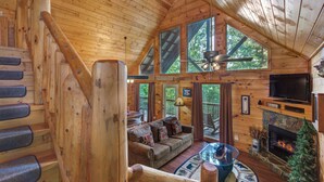 Interior de uma cabana de madeira com escada, sala de estar com lareira e janelas amplas.