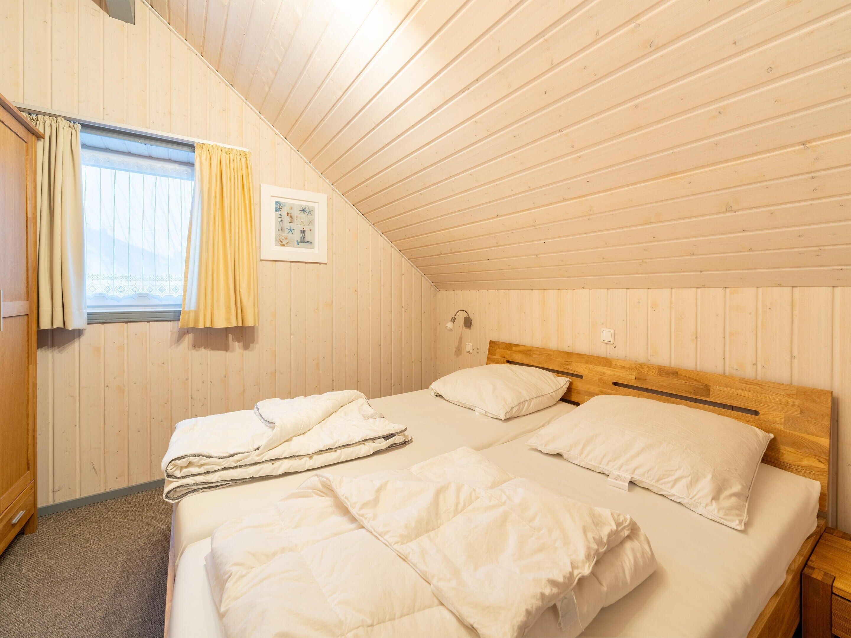 Une cabane confortable, composée d’une seule pièce, avec un plafond en pente, des murs en bois, un lit avec une literie blanche, une petite table de chevet et une fenêtre avec des rideaux.