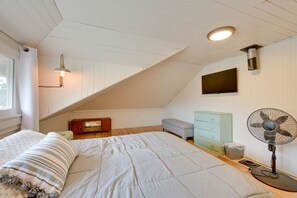 Un dormitorio con techo inclinado, una cama, un aparador, un televisor y un ventilador de pie.
