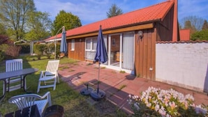 En etagers bygning med rødt tegltag, en overdækket patio med udendørs møbler, og en have med blomster.