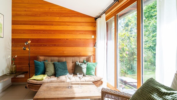 Una stanza accogliente con soffitto in legno, un'ampia finestra, un divano con cuscini, un tavolo con bicchieri e una sedia in vimini.