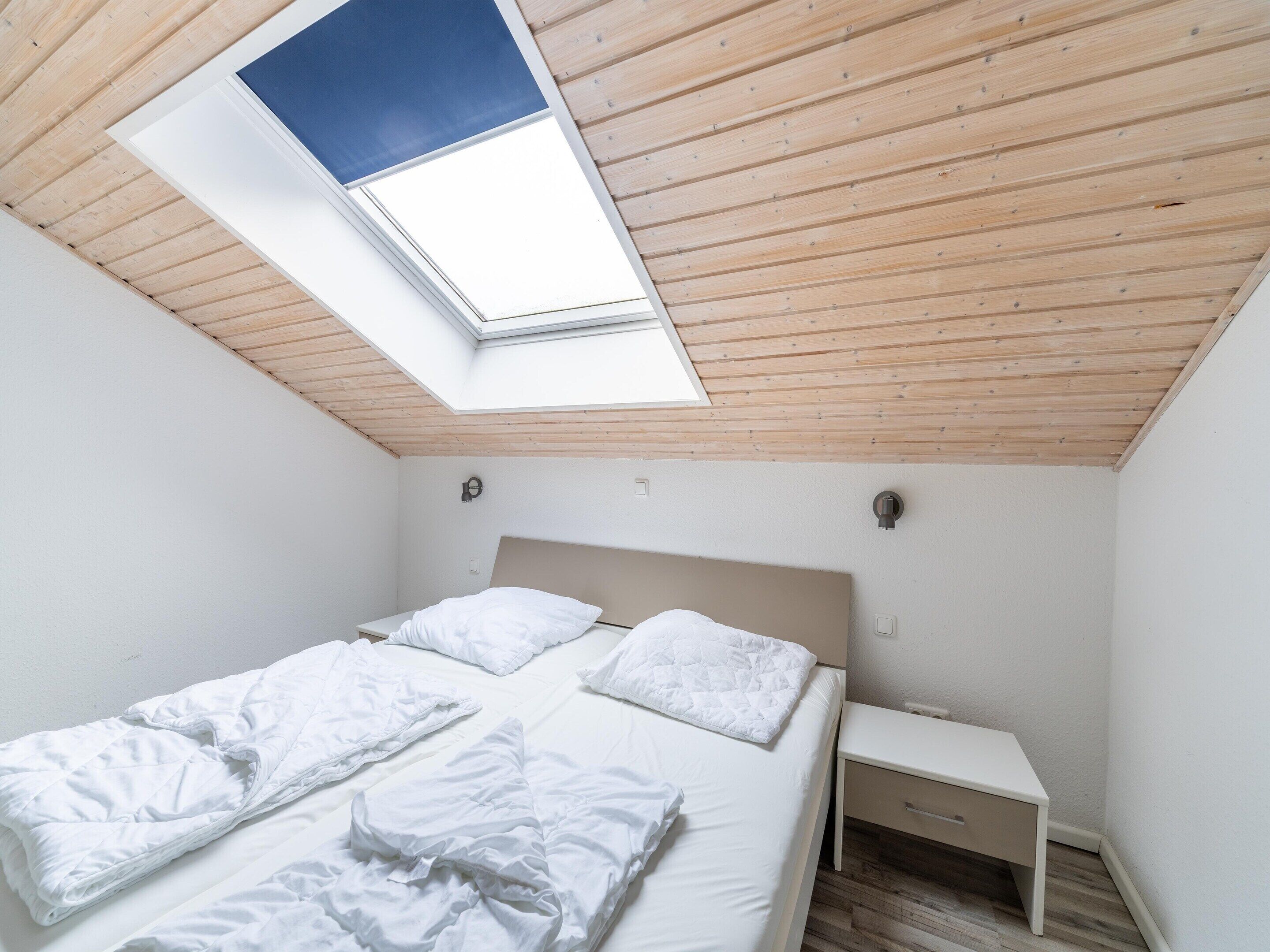 Une chambre avec un puits de lumière, un lit avec une literie blanche, une table de chevet et une petite commode.
