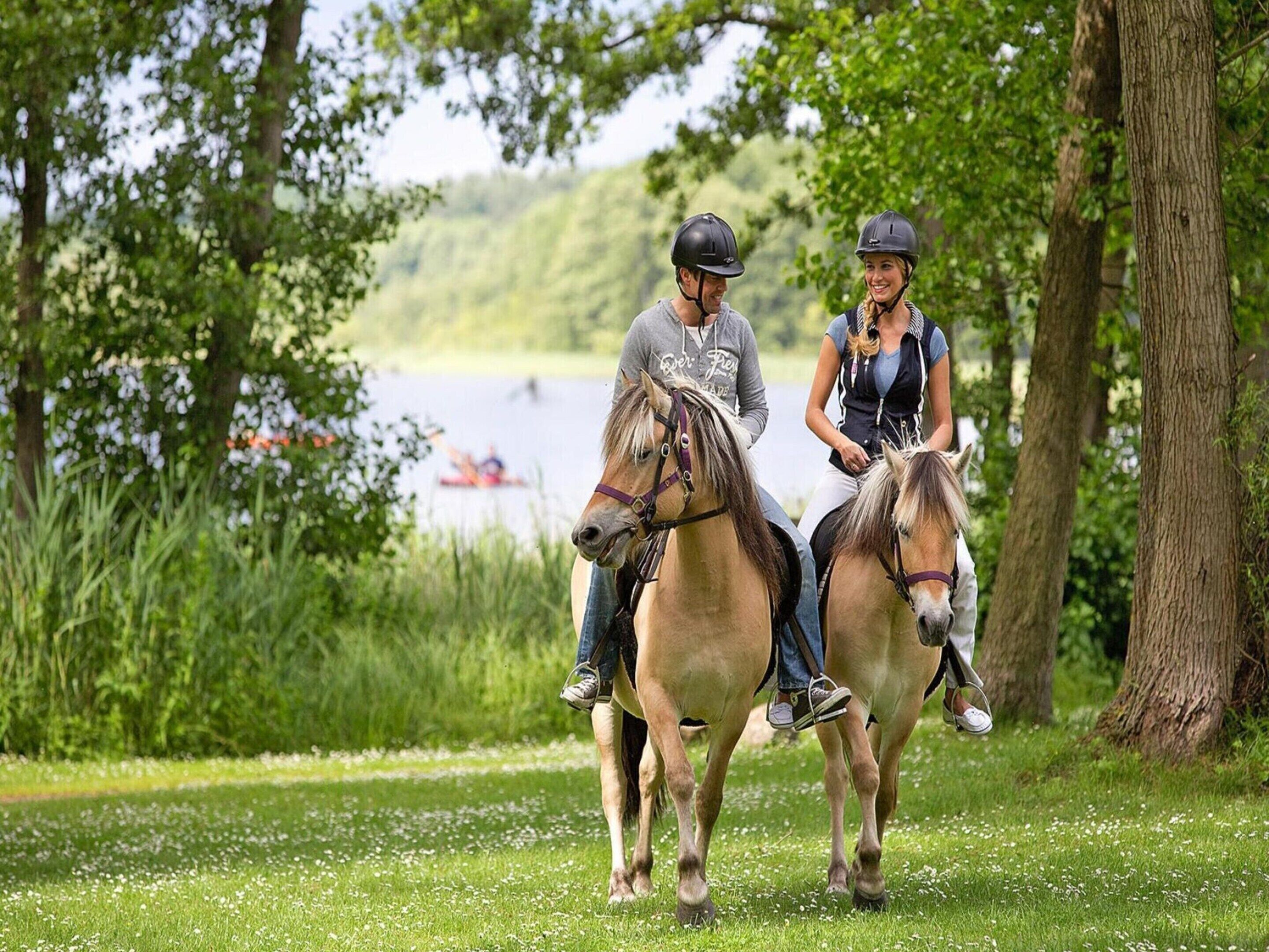 Dos personas montando a caballo por un sendero cubierto de césped, con árboles y un lago al fondo.