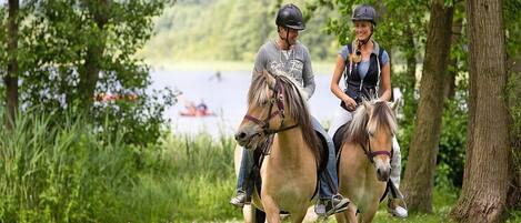 Dos personas montando a caballo por un sendero cubierto de césped, con árboles y un lago al fondo.