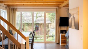 Um quarto espaçoso com escada de madeira, área de jantar com mesa e cadeiras, televisão fixada na parede e porta de vidro deslizante que dá para área externa com vista para árvores e um edifício de tijolos.