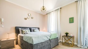 A modern bedroom with a grey upholstered bed, a bedside table with a lamp, a small side table with a plant, and a wall-mounted mirror. - Apartment Bad Camberg (Bad Camberg)
