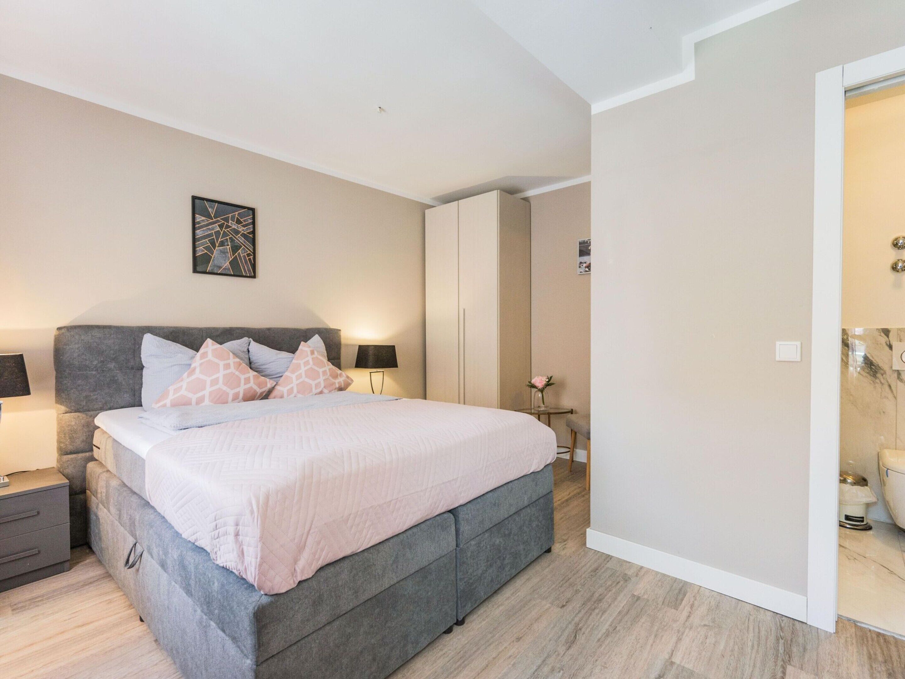 A modern bedroom with a grey upholstered bed, a bedside table with a lamp, a wall-mounted abstract art piece, and a door leading to a bathroom.