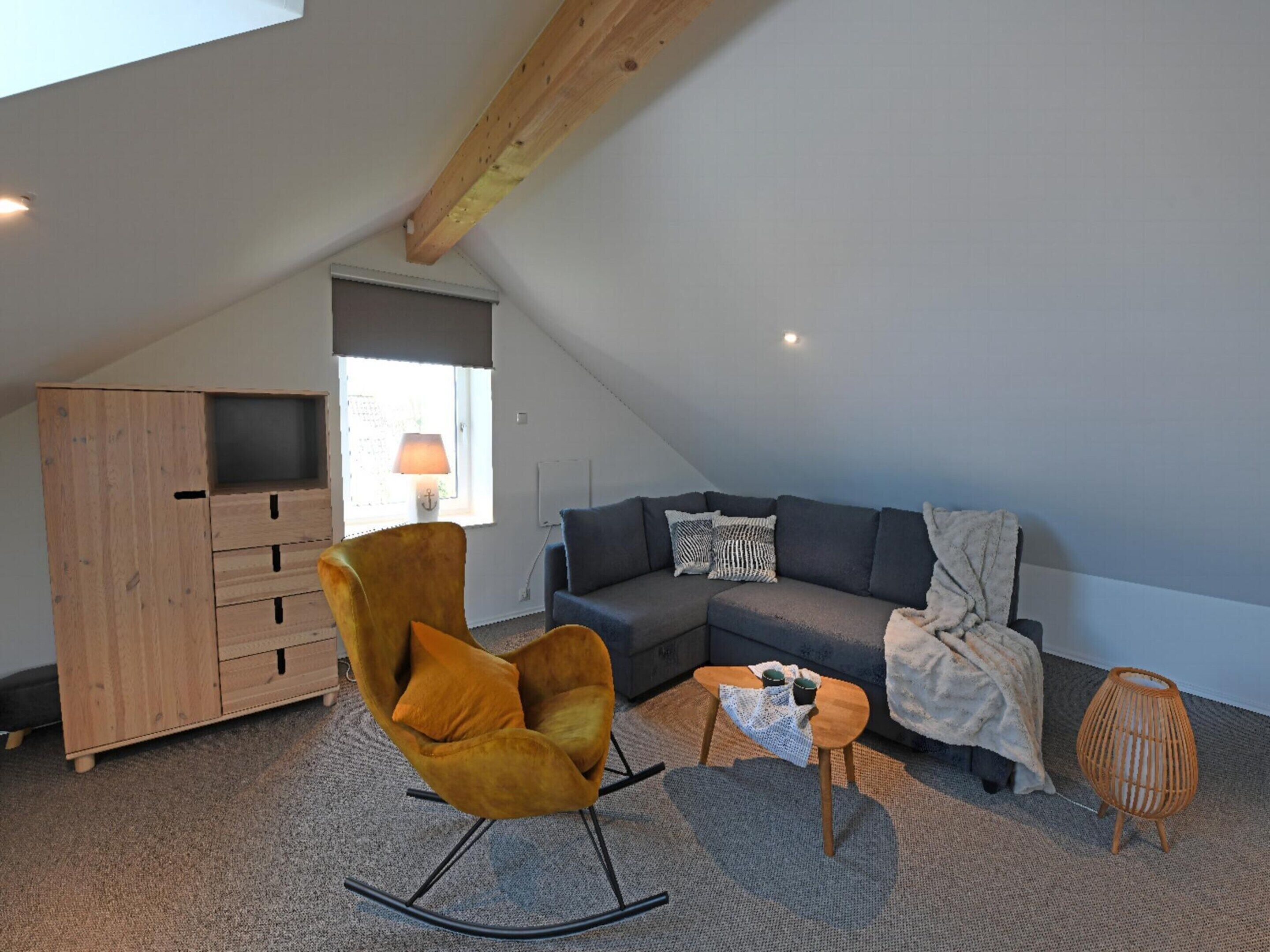 A cozy loft bedroom with a sloped ceiling, a wooden wardrobe, a rocking chair, a grey sofa, and a small table with two cups.