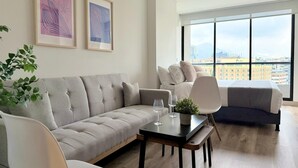 A modern living room with a grey sofa, a dining table with wine glasses, a white chair, and a large window offering a city view. - Urban Chico by Weeki (Bogotá)