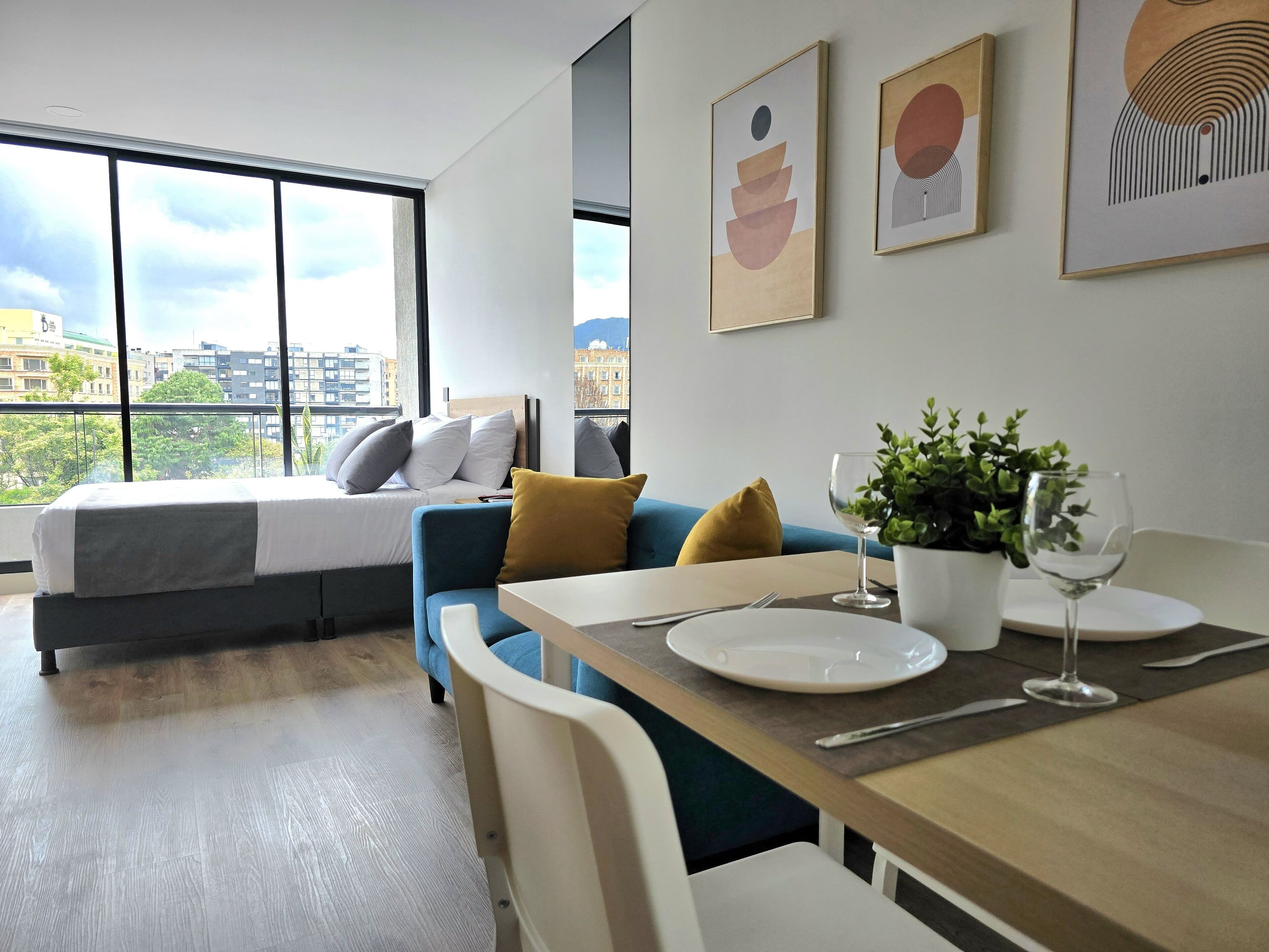 Une chambre d’hôtel moderne équipée d’un lit, d’un canapé, d’une table à manger avec des assiettes et un verre à vin, et d’une grande fenêtre donnant sur la ville.