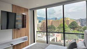 A modern bedroom with a large window, a flat-screen TV mounted on the wall, and a bed with grey bedding. - Urban Chico by Weeki (Bogotá)