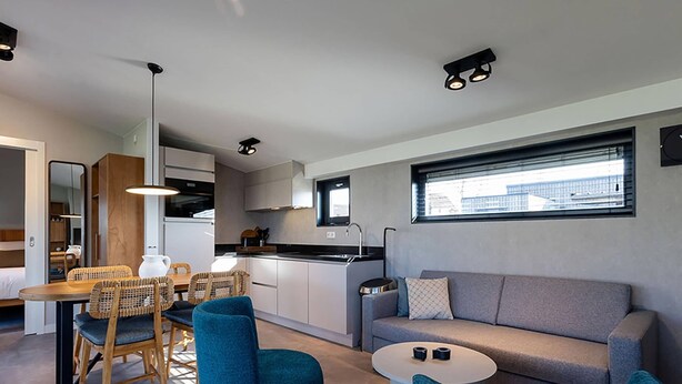 A modern living room with a sofa, two blue chairs, a dining table with wicker chairs, a kitchen area, and a large window providing natural light. - Docklodge at Frisian Lakes With Private Mooring (Delfstrahuizen)