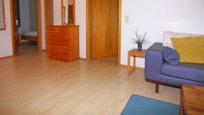 Ein Zimmer mit einem hölzernen Schminktisch, einer Couch mit Kissen, einem kleinen Tisch mit einer Pflanze und einer Tür zu einem anderen Raum.