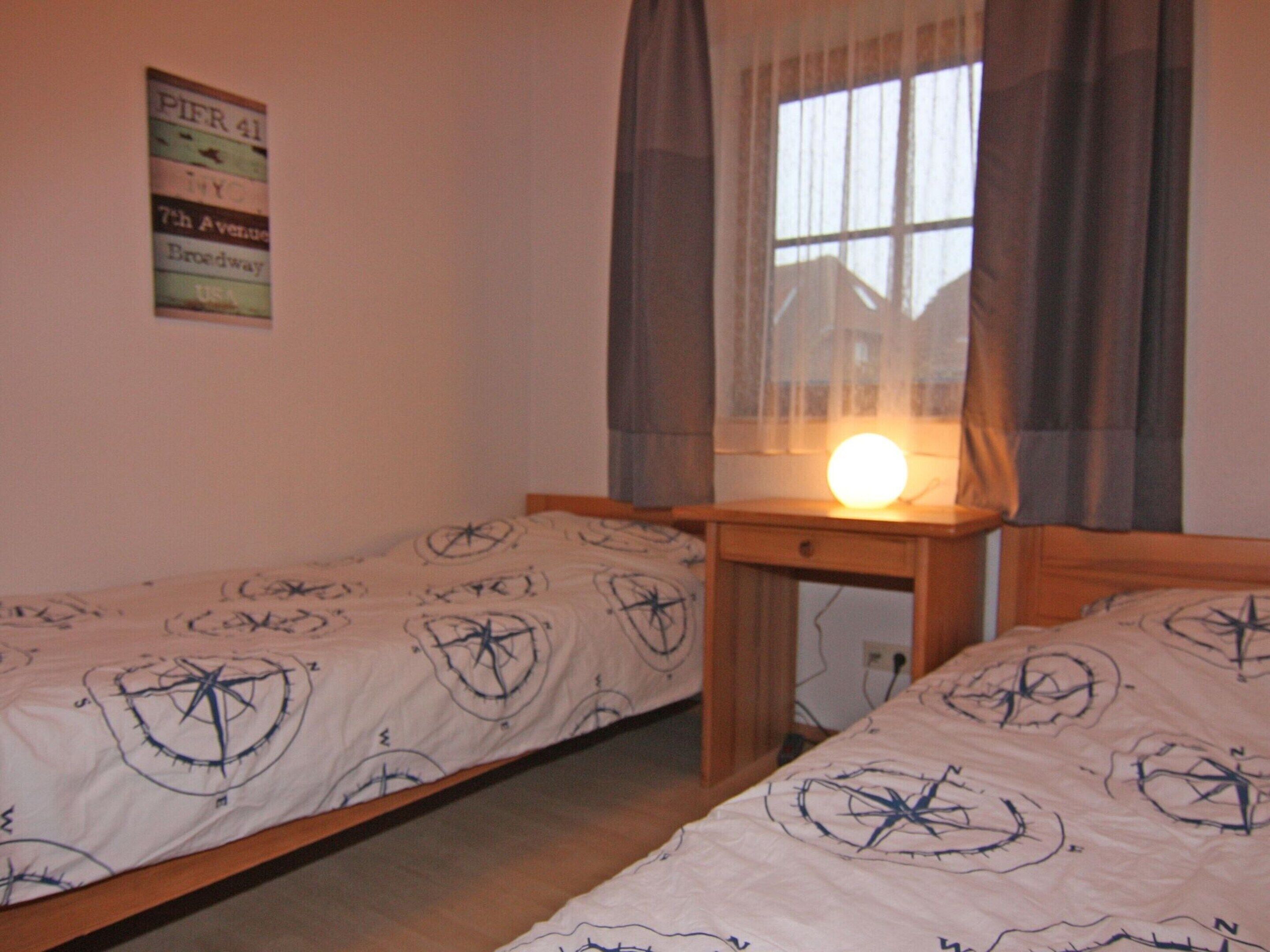 A room with two single beds, a wooden nightstand, a window with curtains, and a wall sign.