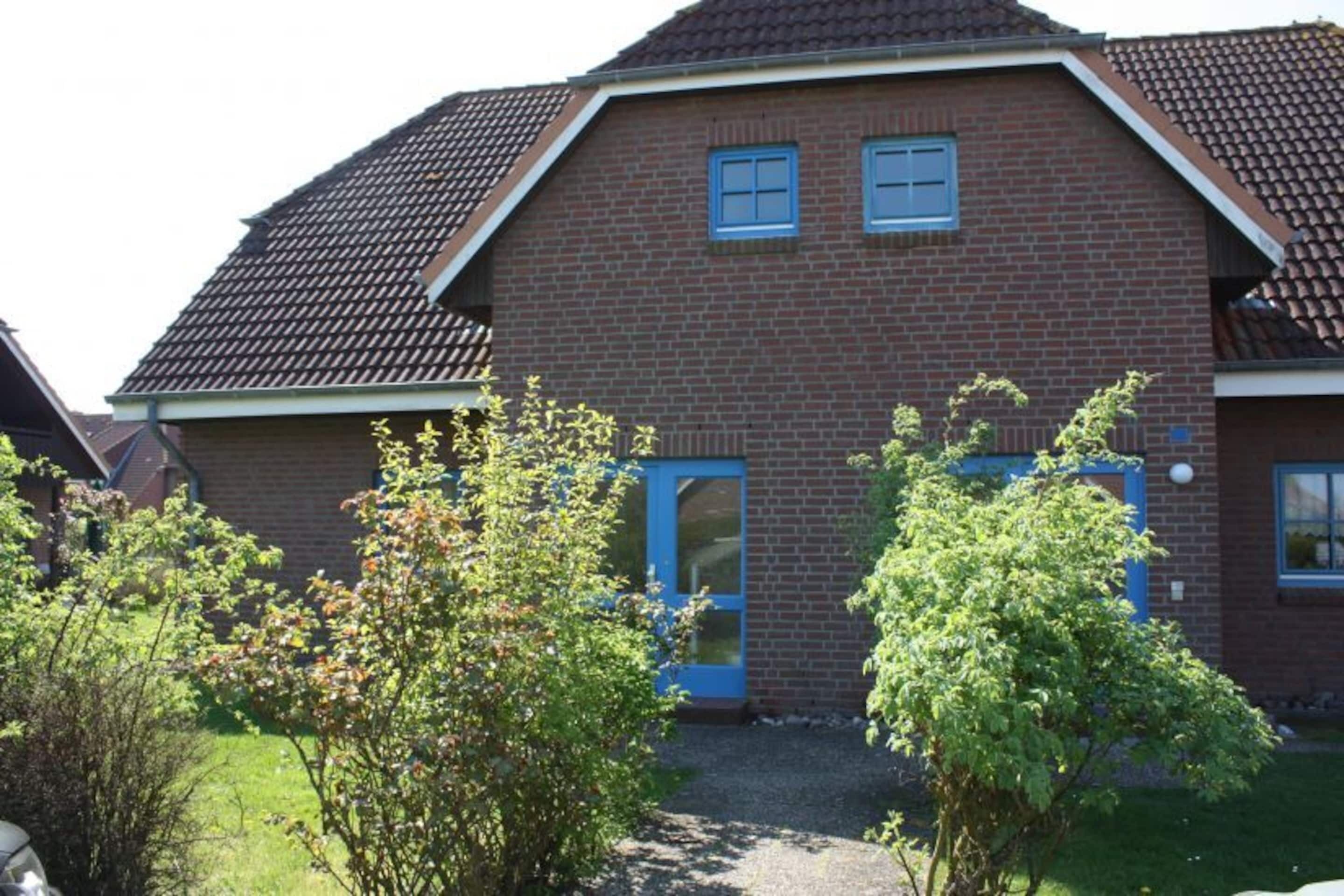 Une maison à deux étages, construite en briques, avec une porte et des fenêtres bleues, entourée de verdure.