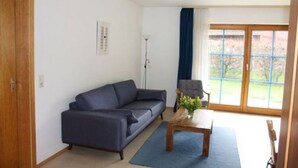 Ein Wohnzimmer mit einem dunkelblauen Sofa, einem Holztisch mit Blumen und einem großen Fenster mit Blick ins Grüne.