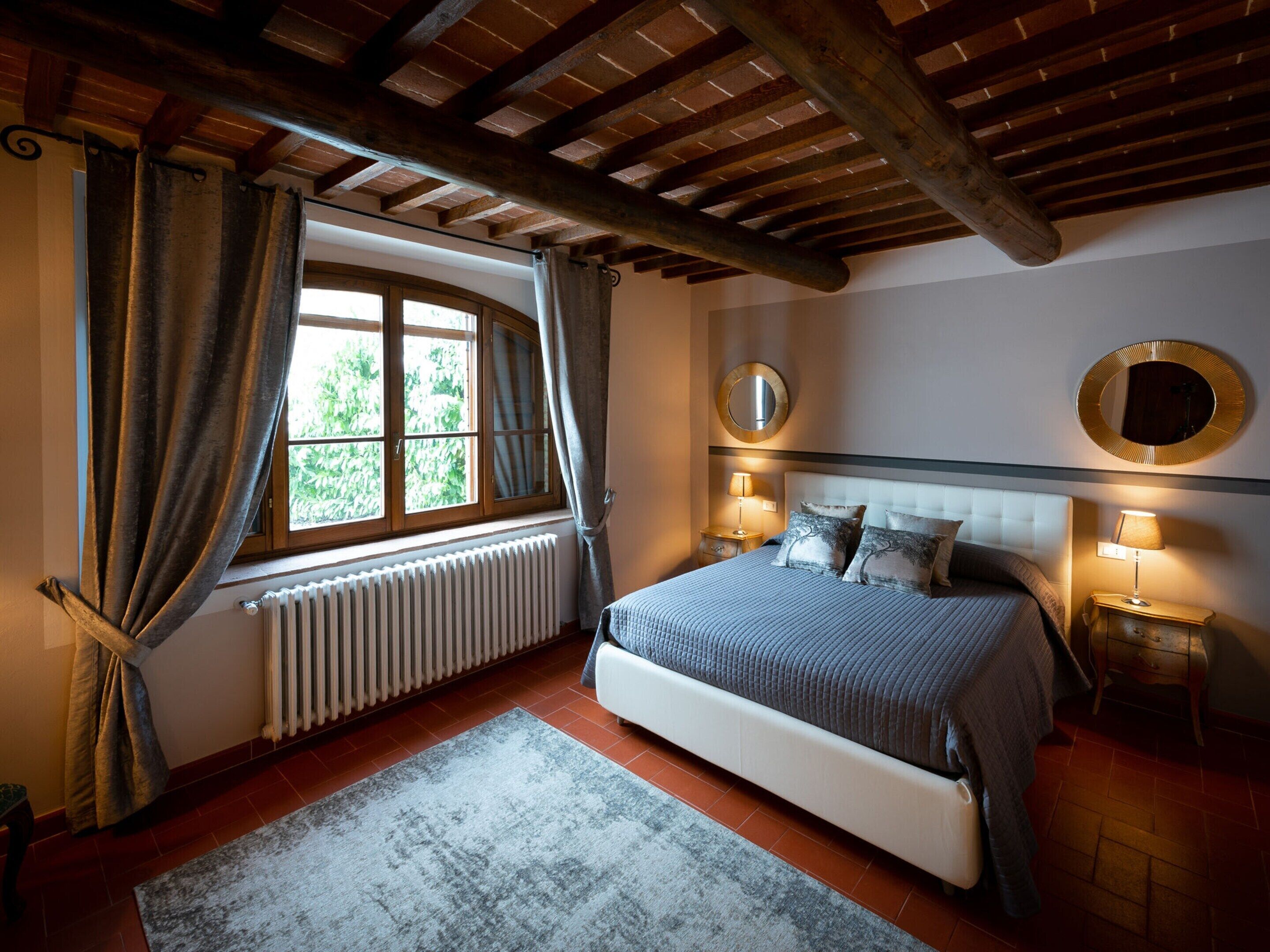 Un dormitorio con una cama grande, dos mesitas de noche con lámparas, una ventana con cortinas, un radiador y un techo de madera.