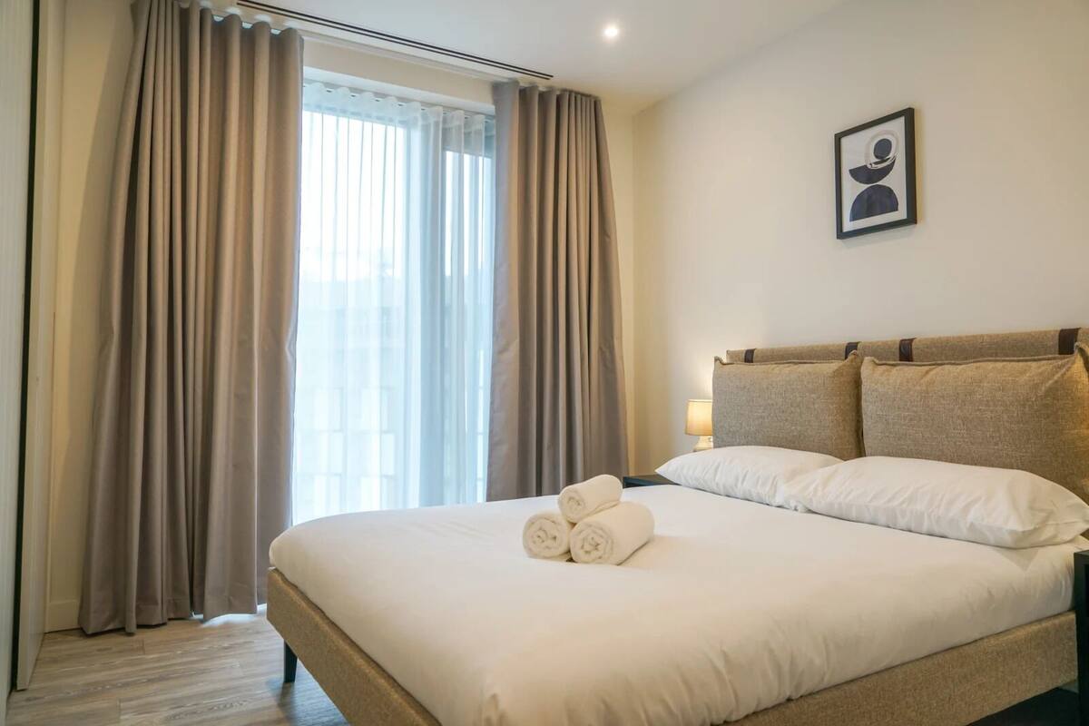 A hotel room with a large bed, beige curtains, a framed picture on the wall, and a neatly made bed with towels.