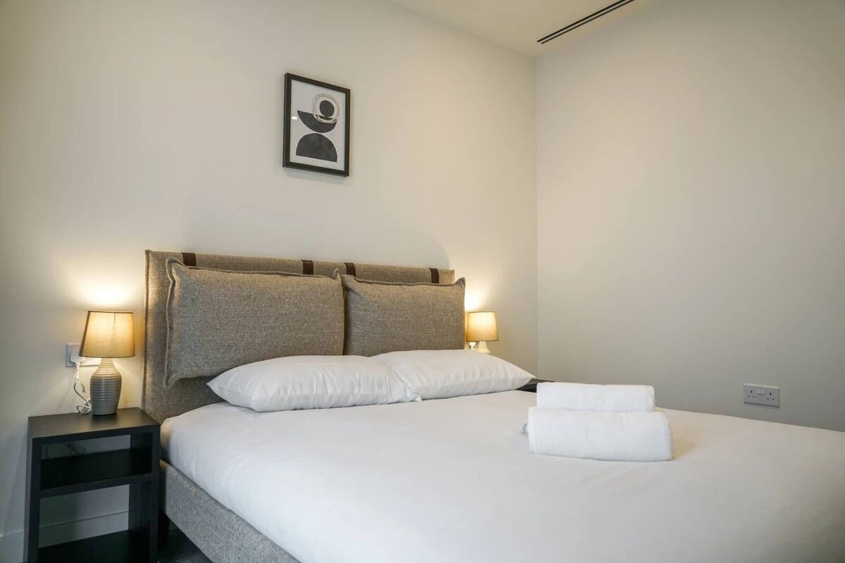 A neatly made bed with white linens and two pillows, a bedside table with a lamp, and a framed picture on the wall.