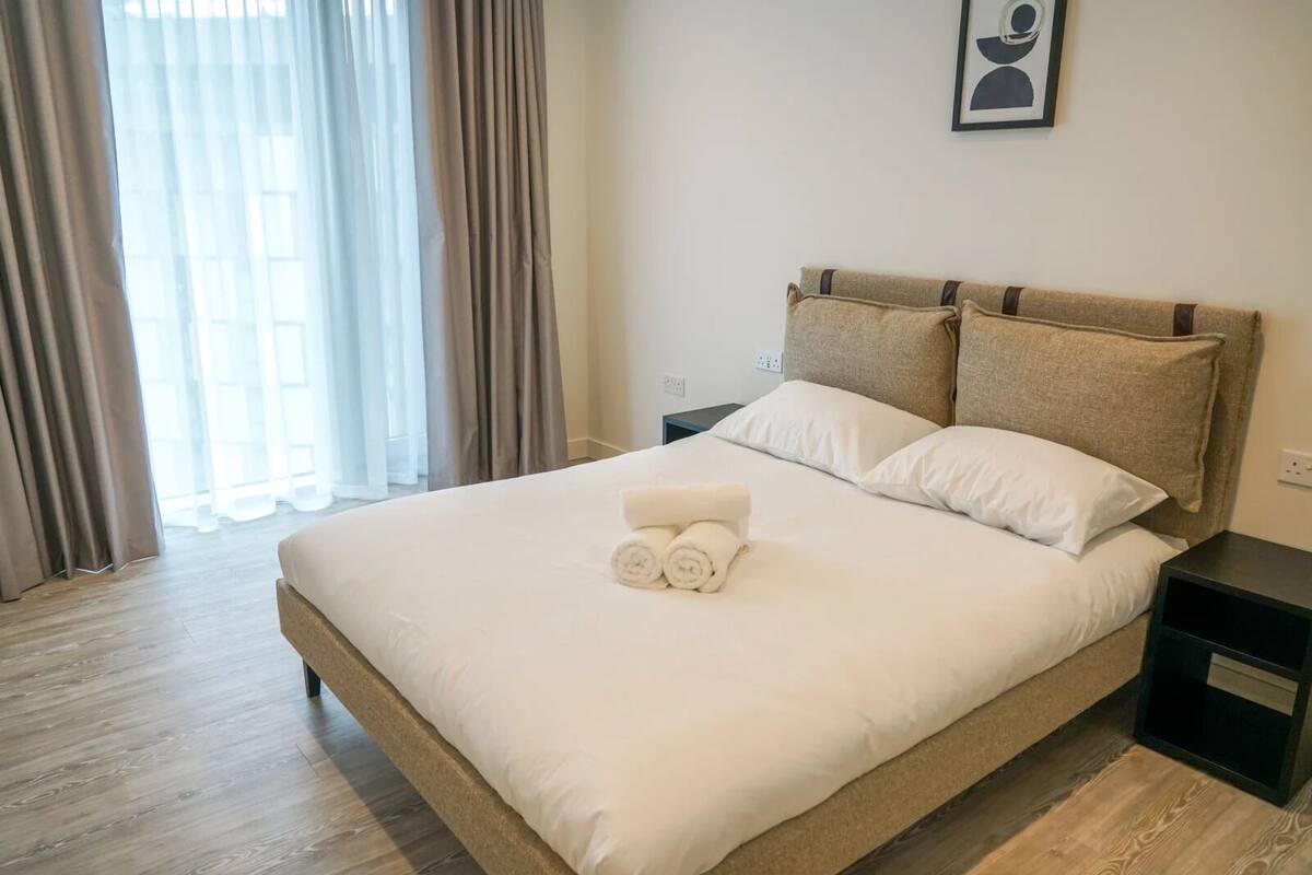 A neatly made bed with white linens and two pillows, a small bedside table, and a window with curtains.