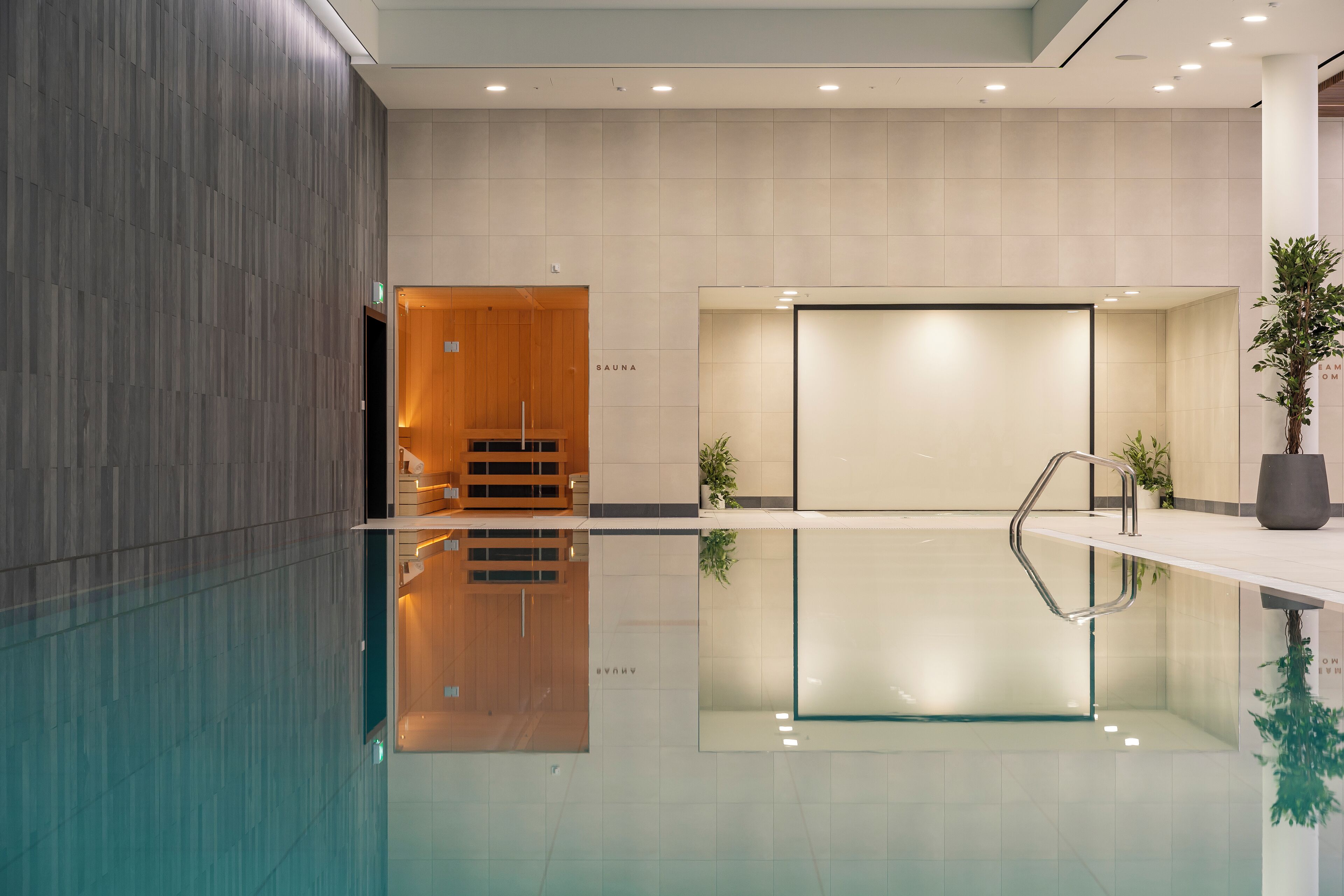 Un espace piscine intérieur moderne, doté d’un sauna en bois et d’un mur de verre qui reflète l’environnement.