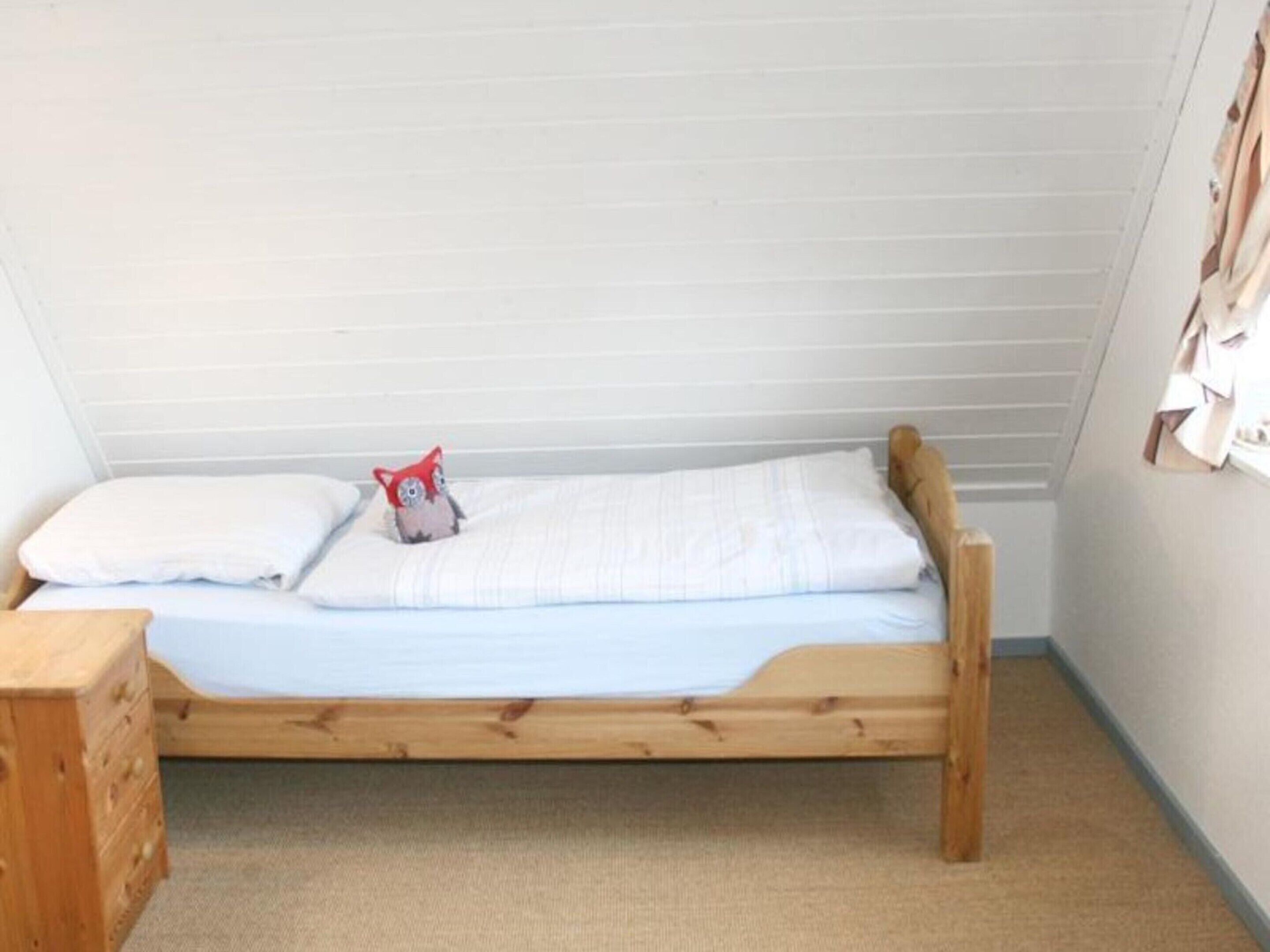 A small, single-bed room with a wooden frame, a white mattress, and a red pillow.
