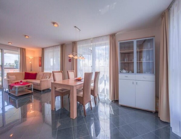A modern living room with a dining area, large windows, and a kitchen with cabinets. - Geschmackvoll Eingerichtetes Appartement (Rechlin)