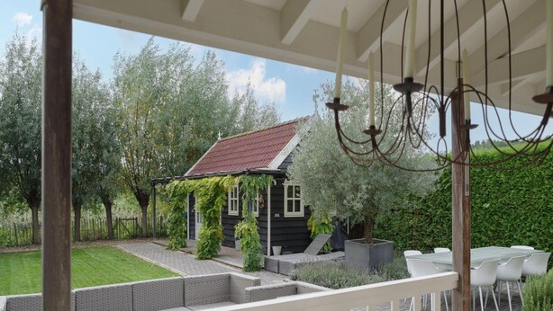 A covered outdoor area with a patio, wicker furniture, a small house with ivy, and a hedge in the background. - Zeeland Retreat; Sauna and Seaside Charm (Wemeldinge)