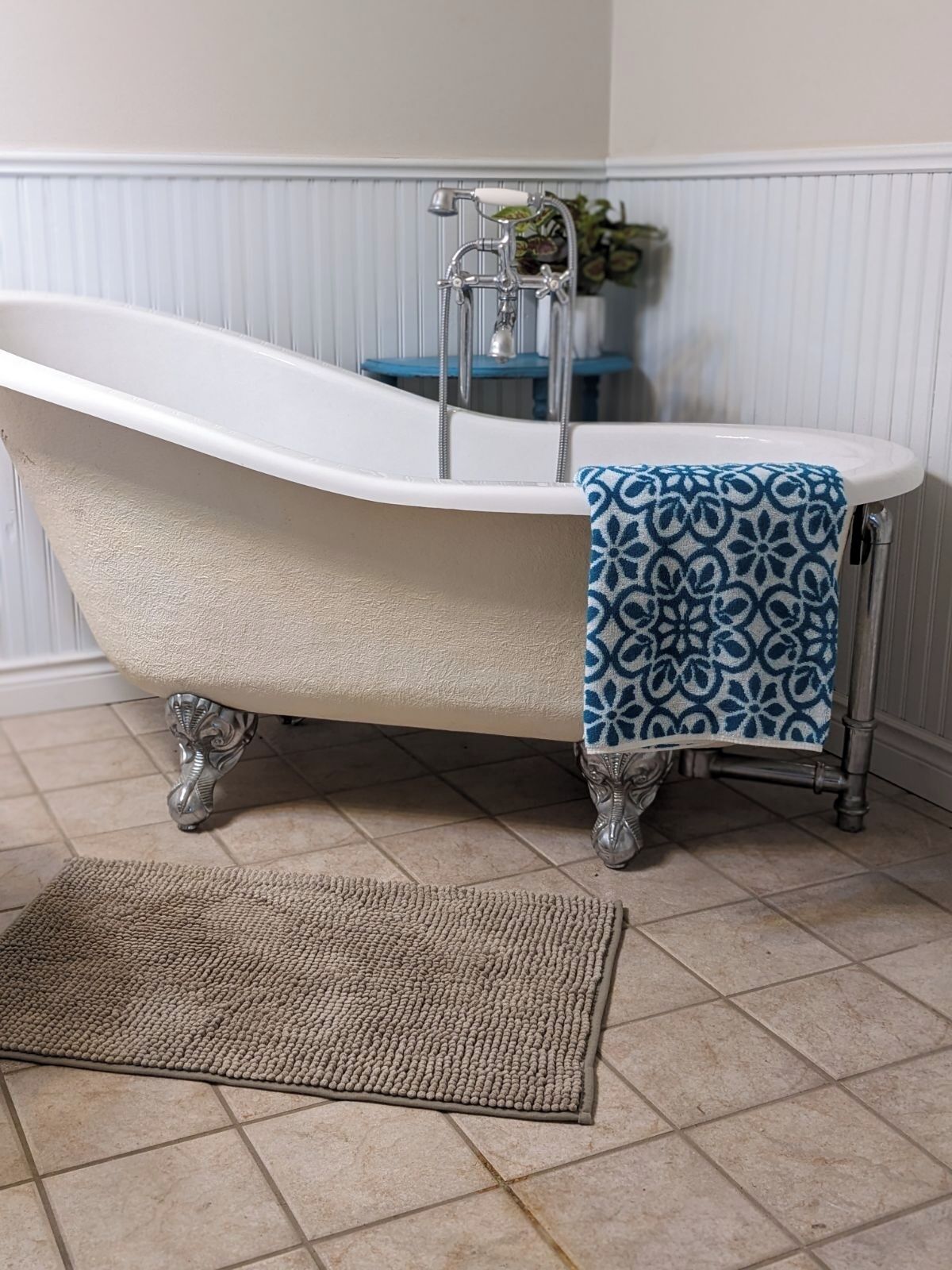 A clawfoot bathtub with a blue patterned towel, a wooden stool, and a textured rug in a bathroom with white paneled walls and a tiled floor.