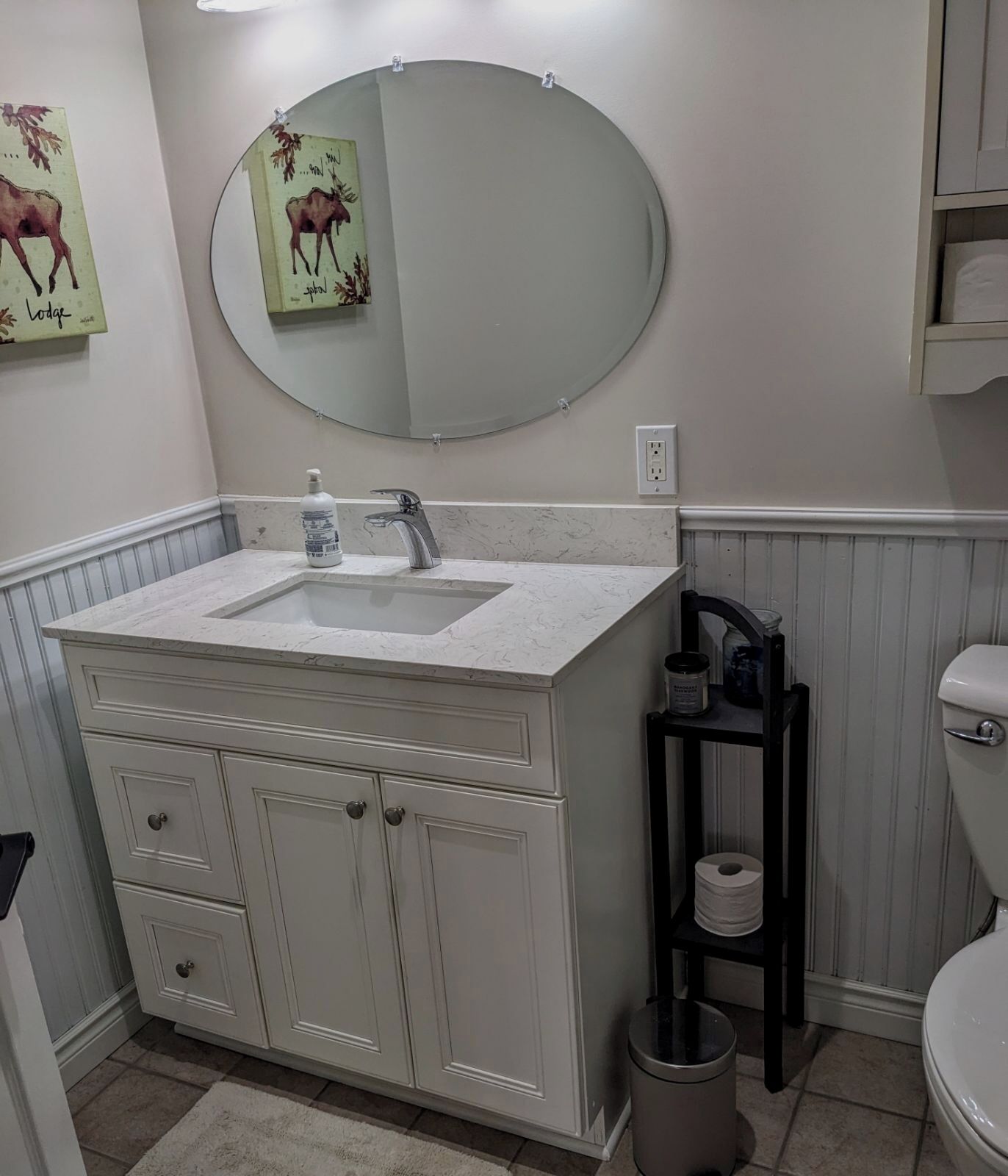 Une salle de bain avec un meuble-lavabo blanc, un miroir rond, des toilettes et une étagère contenant des articles de toilette.
