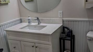 Une salle de bain avec un meuble-lavabo blanc, un miroir rond, des toilettes et une étagère contenant des articles de toilette.