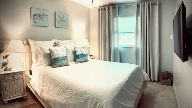 A neatly made bed with white bedding and two decorative pillows, a bedside lamp, and a small nightstand with a lamp. - 01 Coastal Cute 2 Bdrm - FLL-DT-Beach (Fort Lauderdale)