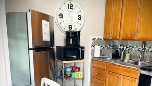 A compact kitchen with wooden cabinets, a microwave, a clock, and a dining area with white chairs and plates. - 01 Coastal Cute 2 Bdrm - FLL-DT-Beach (Fort Lauderdale)