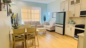 Un salon moderne comprenant un canapé, une table à manger avec des chaises, une cuisine avec un réfrigérateur et une cuisinière, et une grande fenêtre avec des stores.