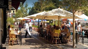Un café en plein air avec des chaises et des tables en osier, abritées par de grands parasols, et entouré d’arbres et d’immeubles.
