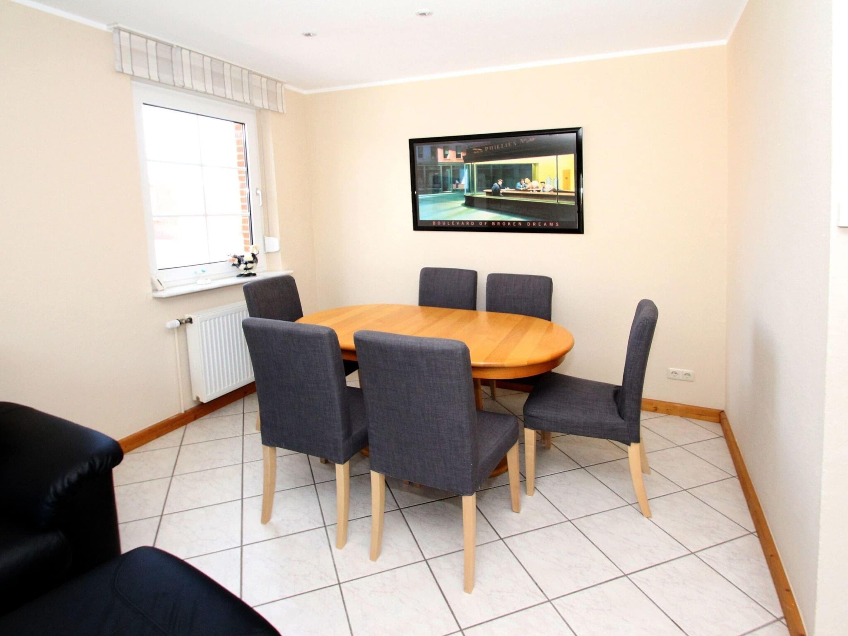 A conference room with a round wooden table, six grey upholstered chairs, a large framed picture on the wall, a window with blinds, and a radiator.