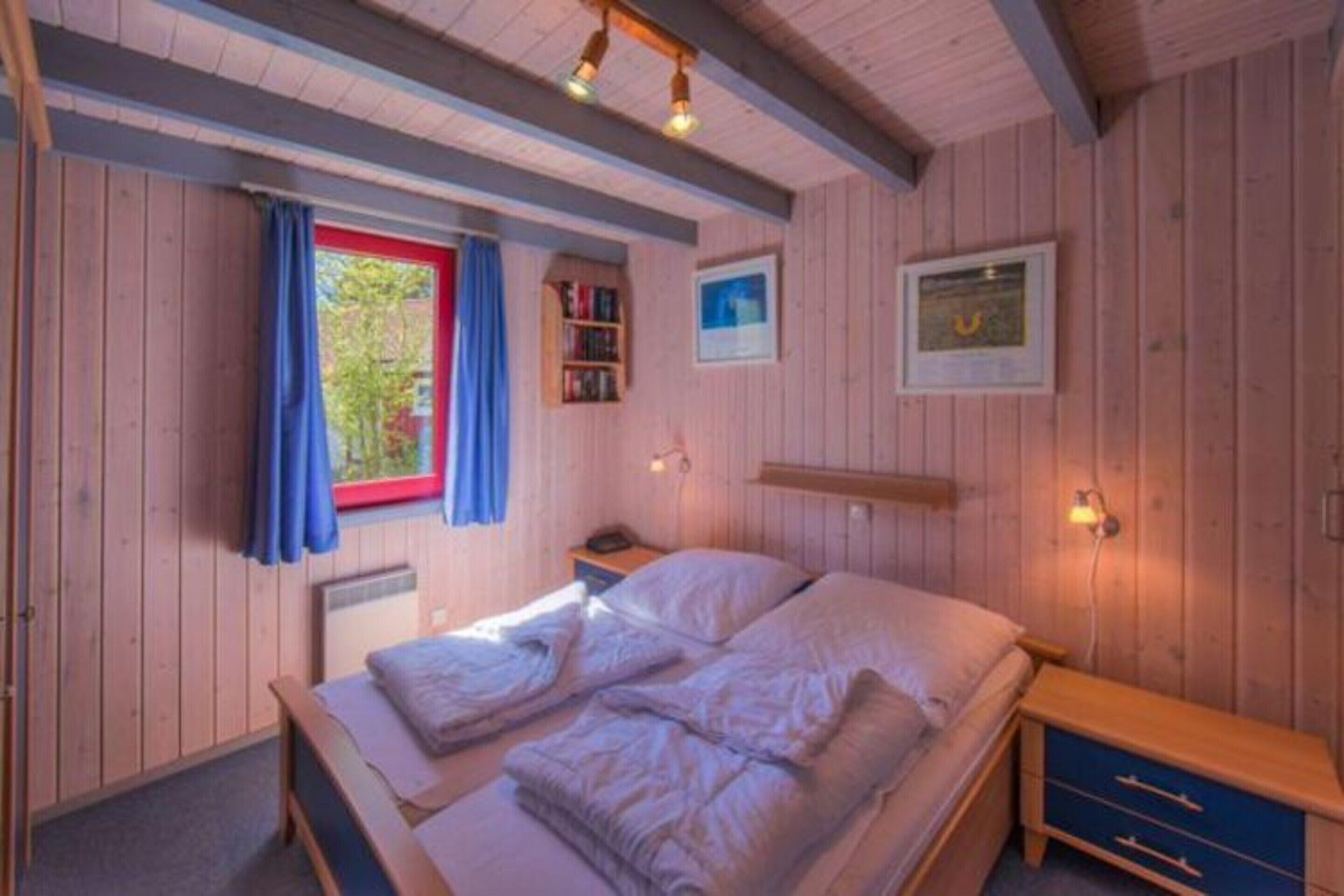 A cozy bedroom with a wooden ceiling, a single bed, a nightstand, a shelf with books, and a window with curtains.