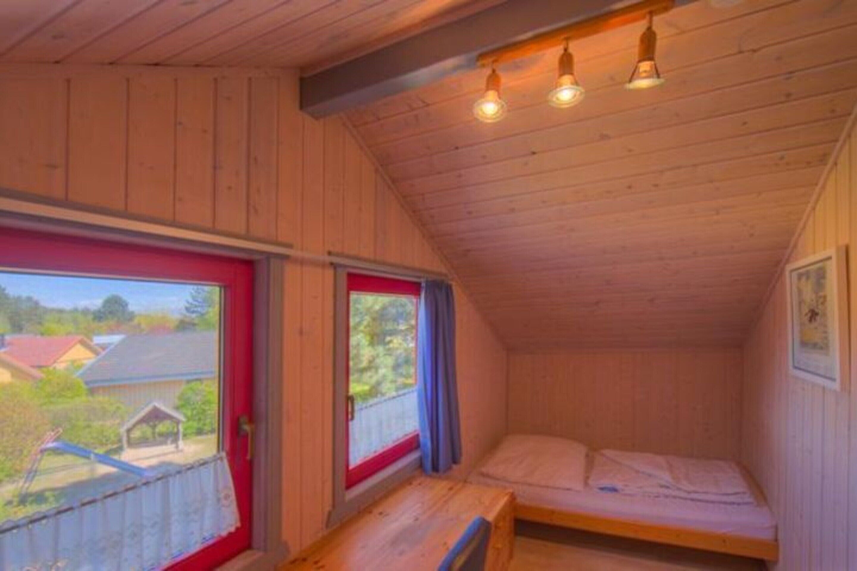 A small cabin with a single bed, a bench, and a window overlooking a house.