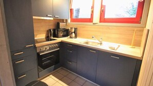 A compact kitchen with dark cabinets, a stove, a sink, and a window with red curtains. - Gemutliches Ferienhaus im Skandinavischen Stil (Mirow)