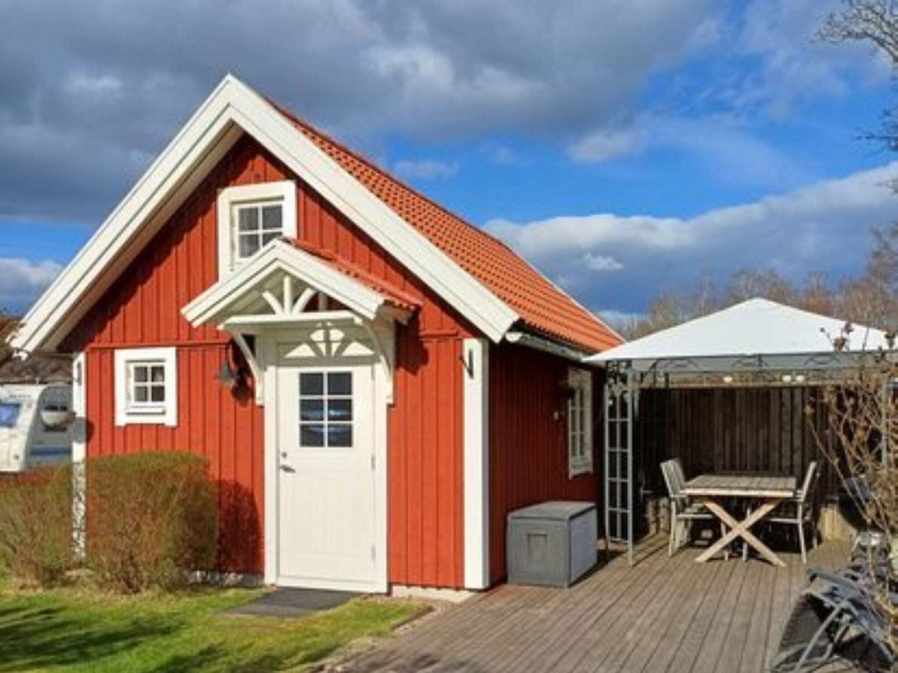 A red wooden house with a white door and windows, a covered porch with a table and chairs, and a small shed.