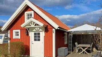 A red wooden house with a white door and windows, a covered porch with a table and chairs, and a small shed.