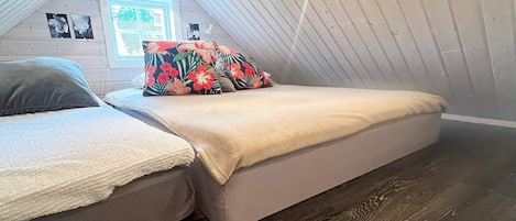 Een kleine, gezellige kamer met een schuine kap, een bed met een bloemenkussen en een raam met uitzicht op groen.