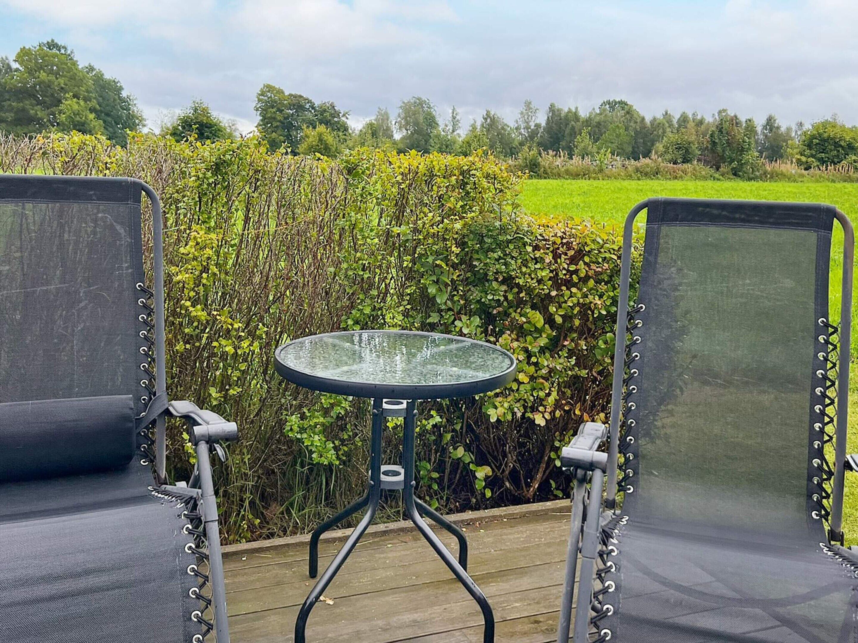 A deck with two chairs and a small table, overlooking a green field and dense bushes.