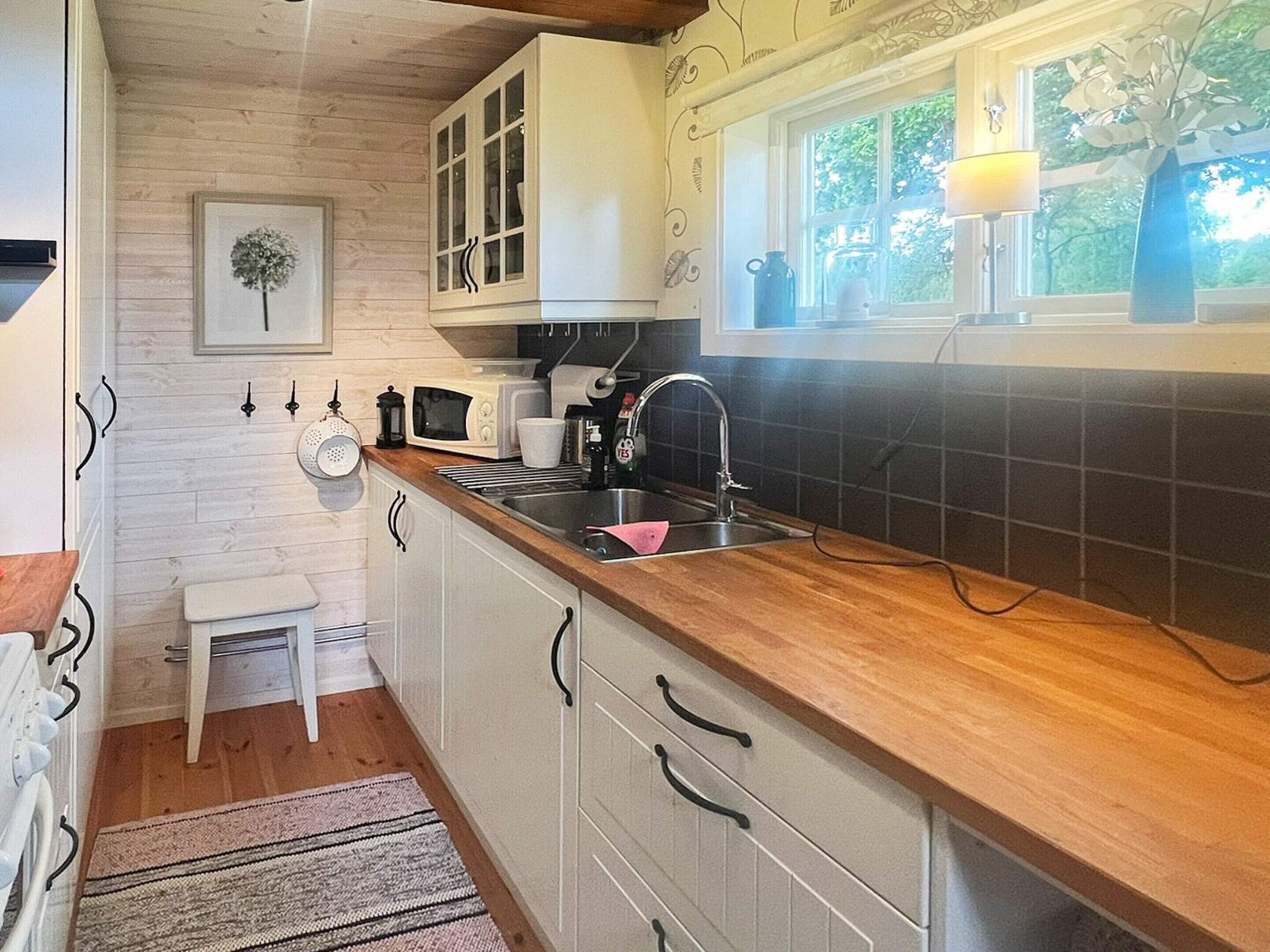 A compact kitchen with white cabinets, a wooden countertop, a sink, a microwave, and a window with a view of greenery.