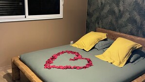 Een bed met een hartvormige decoratie van rozenblaadjes, gele kussens en een groene sprei in een kamer met een raam en behang met een patroon.