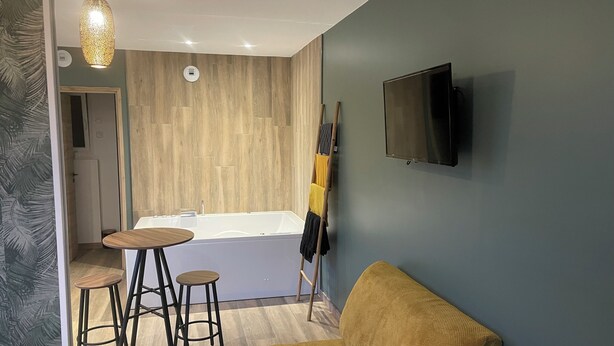 Een moderne badkamer met een badkuip, houten vloeren en een douchegedeelte. Er is een klein tafeltje met krukken, een gele stoel en een tv aan de muur bevestigd.