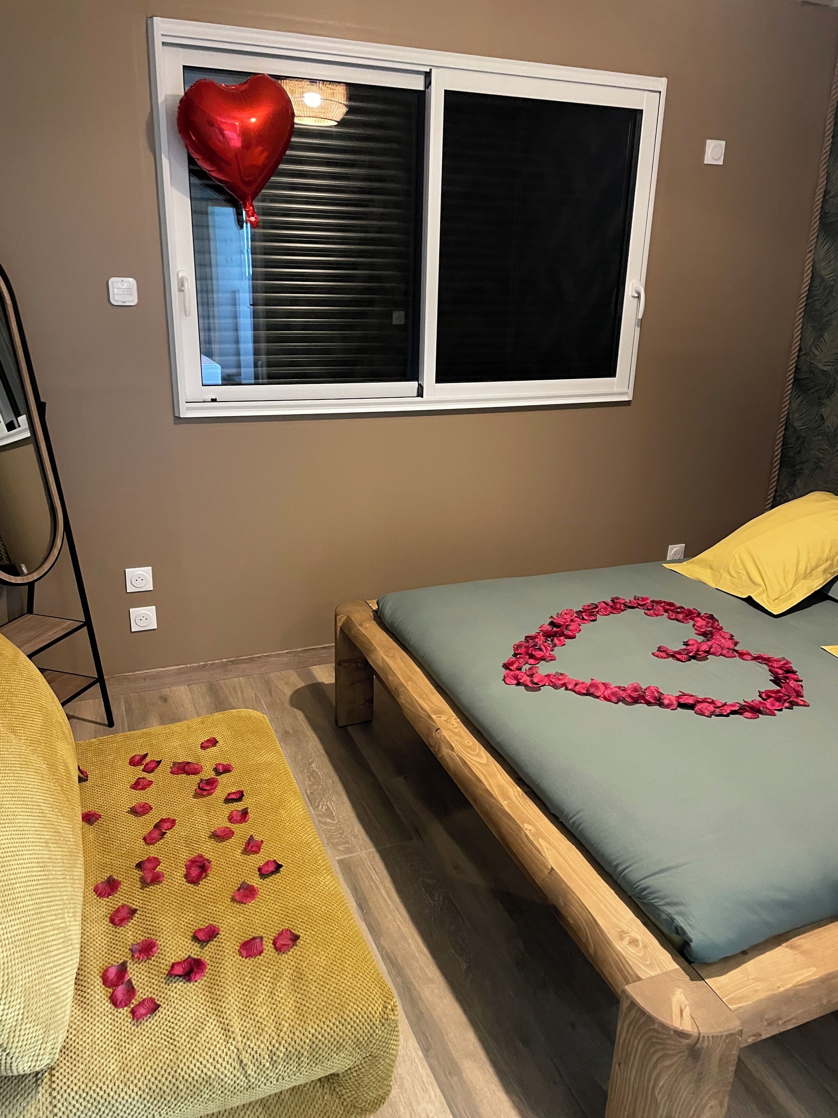 A bedroom with a bed, a window with blinds, a yellow chair, and a heart-shaped decoration on the bed.