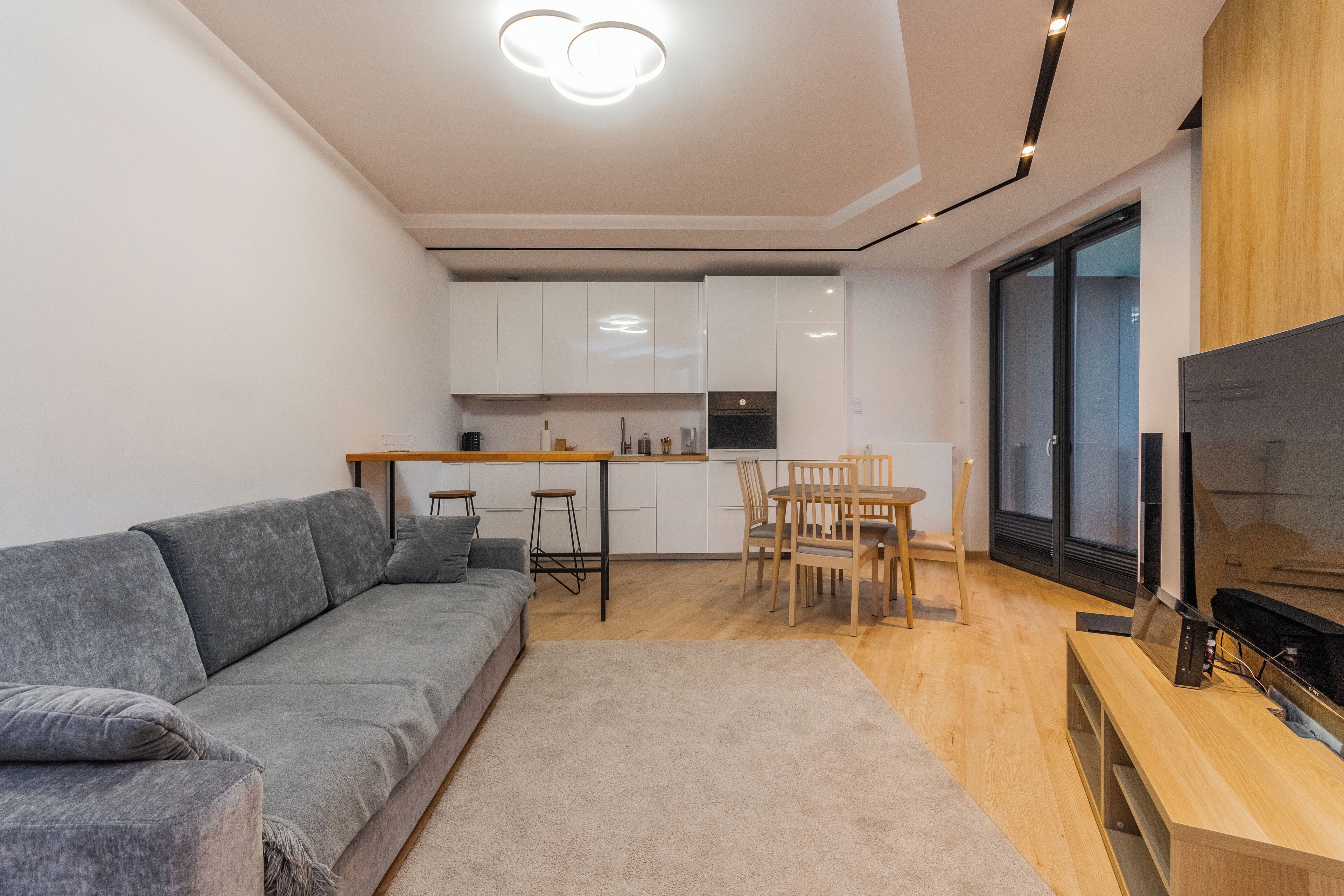 A modern living room with a grey sofa, a wooden dining table with chairs, a TV, and a kitchen area with white cabinets and a wooden countertop.