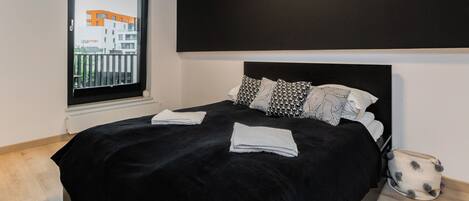 Una habitación de hotel moderna con una cama grande, almohadones blancos y negros estampados, un cabecero negro y vistas a un edificio por la ventana.