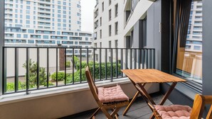 A balcony with a wooden table and two cushioned chairs, overlooking a cityscape with modern buildings.